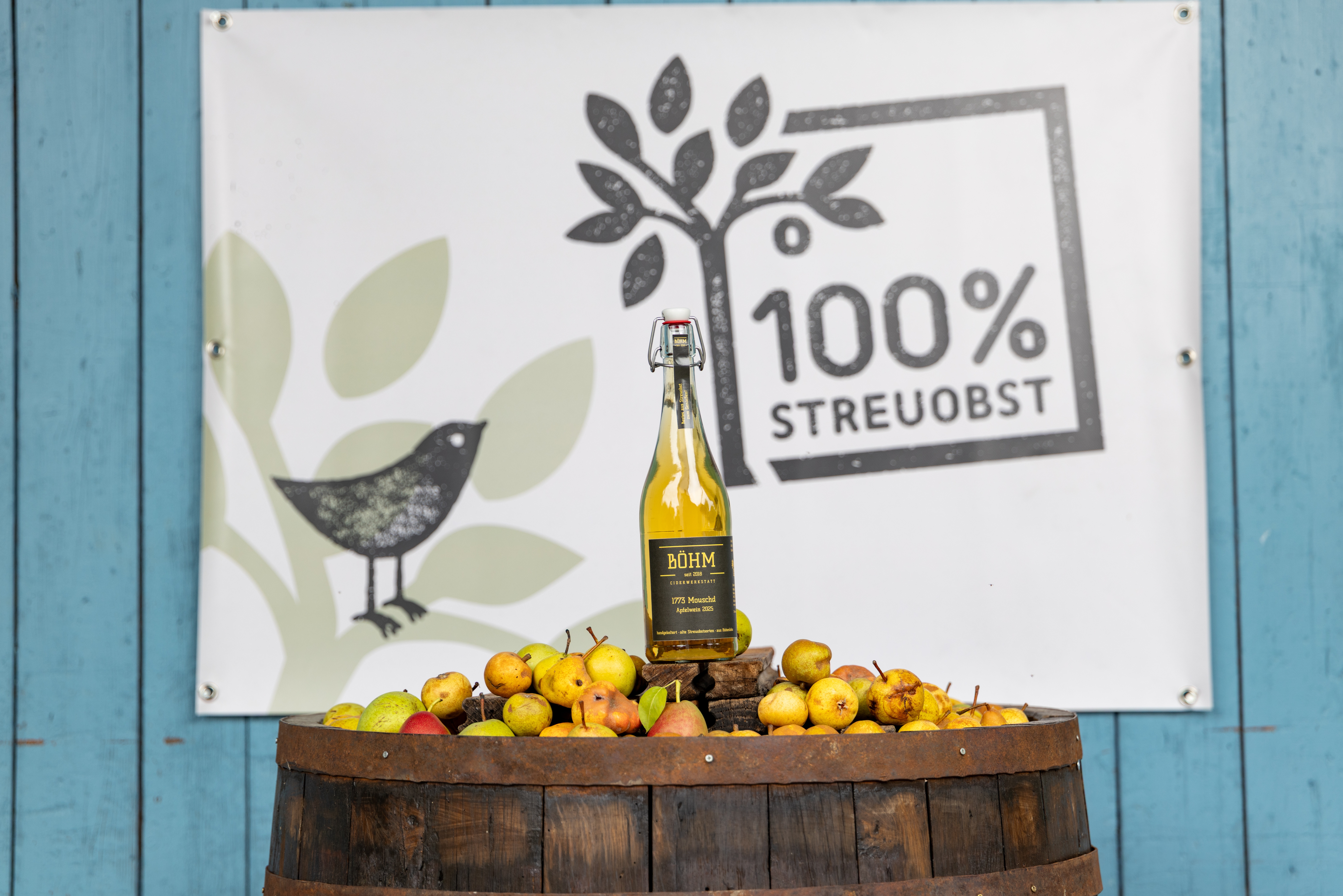 Eine Flasche Cider steht auf einem Fass umgeben von Streuobst, im Hintergrund hängt ein Poster mit dem Logo 100% Streuobst